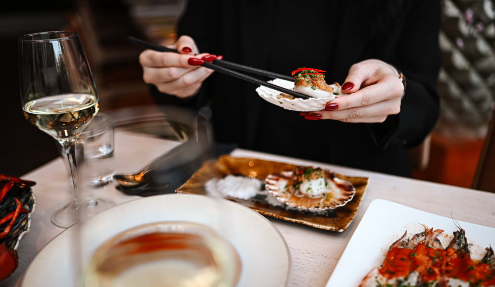 Persoon eet een gerecht uit een schelp met eetstokjes aan een gedekte tafel met wijnglazen Persoon eet een gerecht uit een schelp met eetstokjes aan een gedekte tafel met wijnglazen