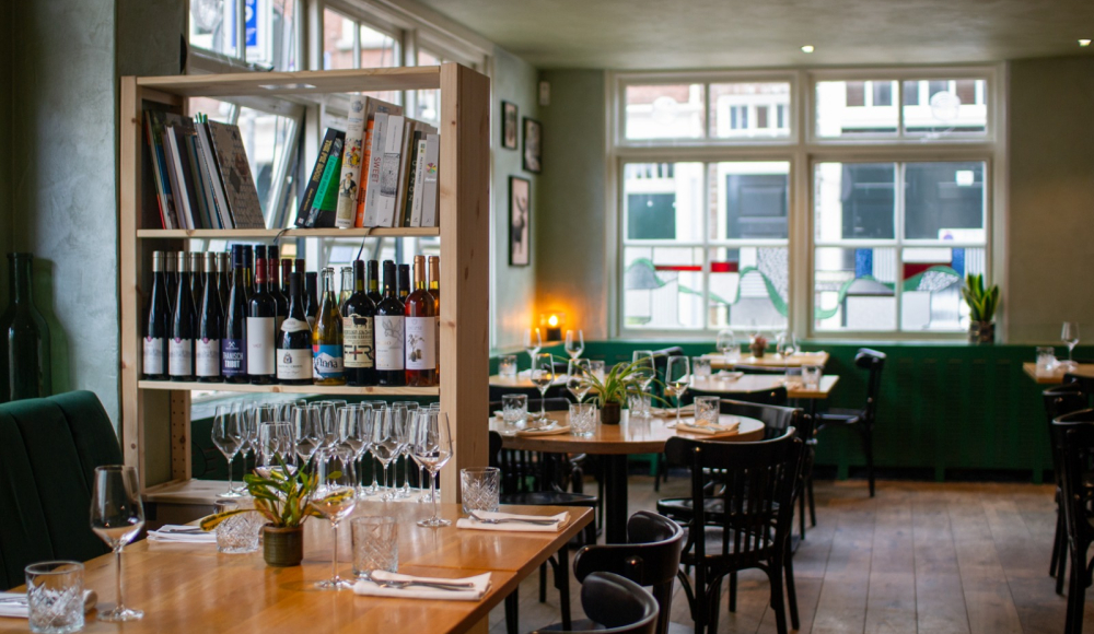 Intérieur chaleureux du restaurant avec tables en bois, chaises noires, tables dressées et étagère ouverte exposant des bouteilles de vin, des verres et des livres de cuisine. Intérieur chaleureux du restaurant avec tables en bois, chaises noires, tables dressées et étagère ouverte exposant des bouteilles de vin, des verres et des livres de cuisine.