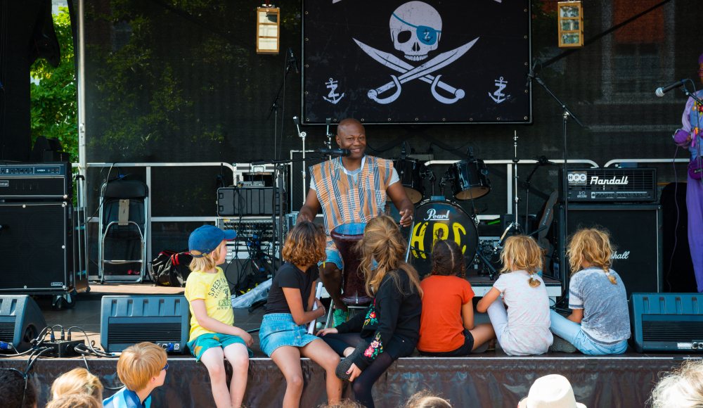 Kinderen zittend voor en op het podium aandachtig luisterend naar artiest tijdens een workshop.
