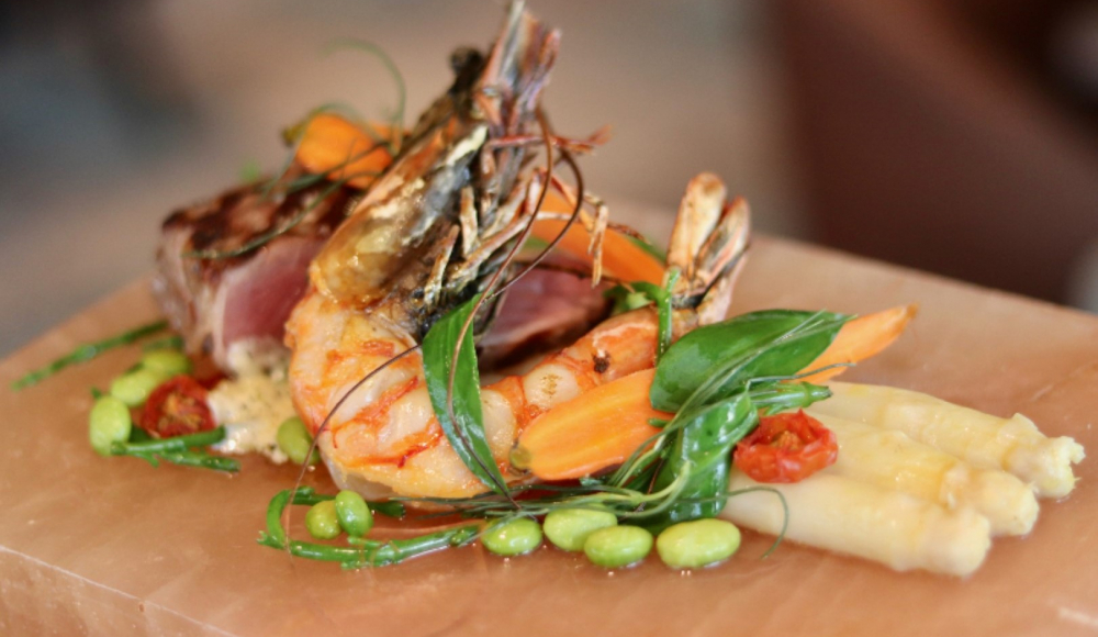 Een houten plank met gegrilde garnaal en kleurrijke garnituur op een bedje van witte asperges bij SALT Coastal Bistro Een houten plank met gegrilde garnaal en kleurrijke garnituur op een bedje van witte asperges bij SALT Coastal Bistro