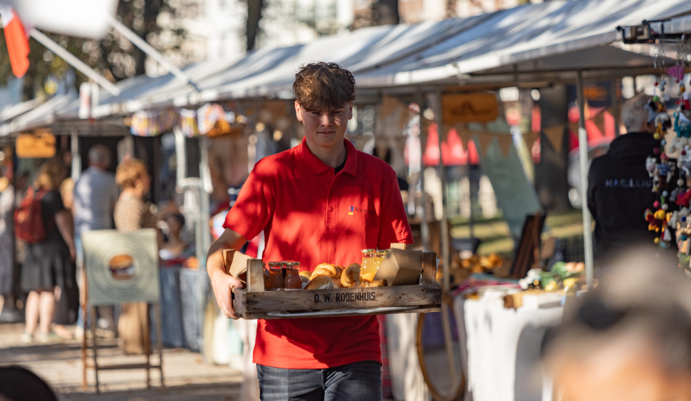 Een jongen in een rode polo draagt een dienblad met eten op een markt.