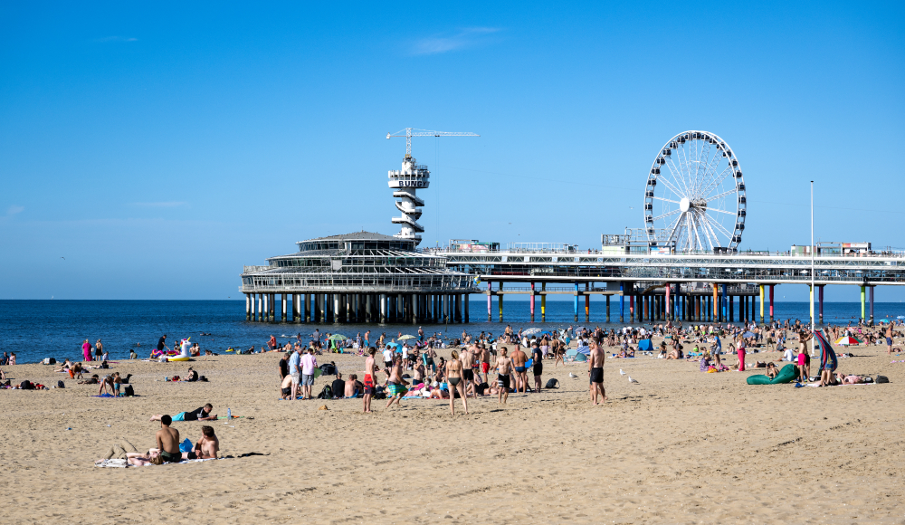 Mensen ontspannen op het strand van Scheveningen met de pier en de reuzenrad op de achtergrond.