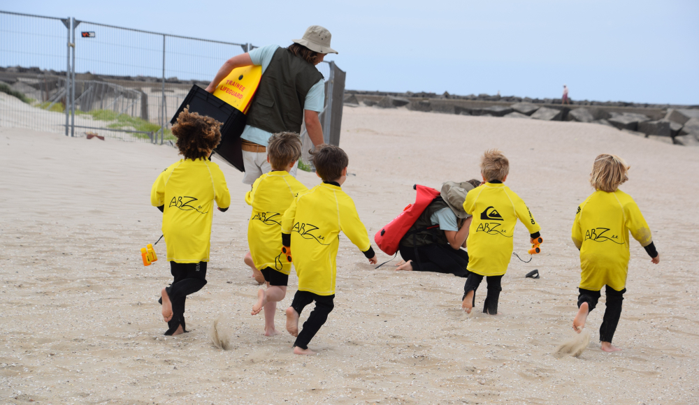 Fünf Kinder in gelben Schwimmshirts rennen am Strand.