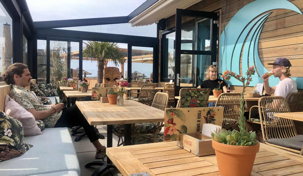 Menschen sitzen an einem Holztisch in einem Café mit Blick auf das Meer.