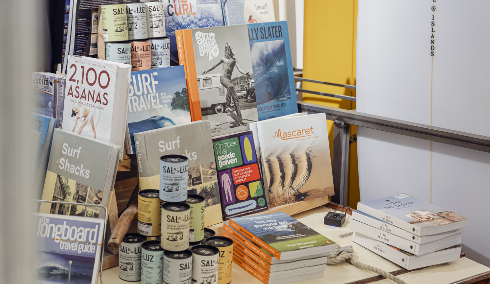 Een tafel met surfboeken, blikken en surfplanken op de achtergrond.