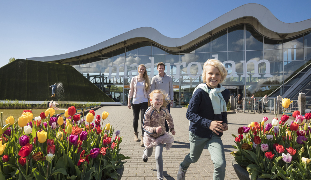 Een vrolijk gezin met twee kinderen loopt tussen kleurrijke tulpen bij de ingang van Madurodam. 
