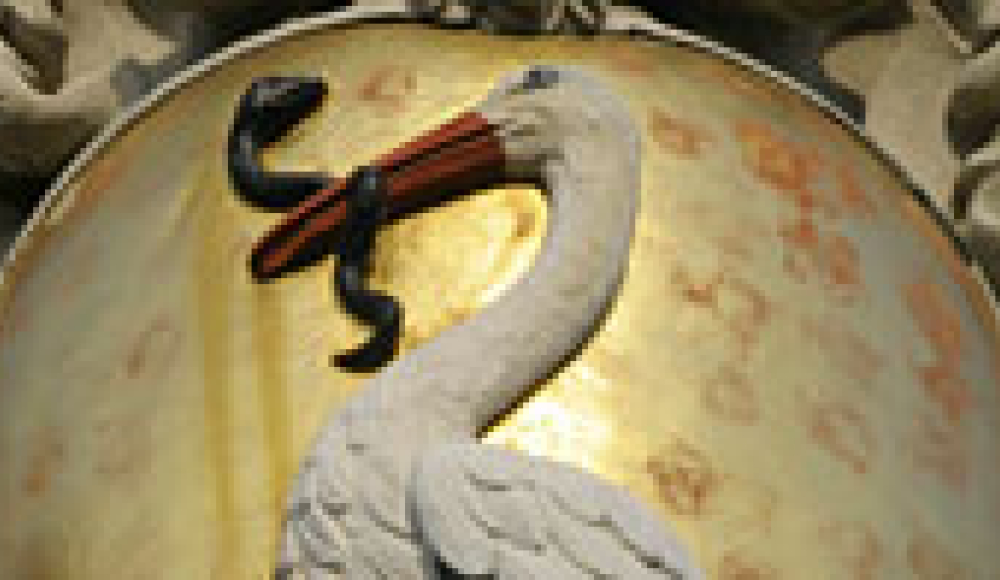 A close-up of the coat of arms of The Hague, featuring a white stork holding an eel in its beak, set against a golden shield background. The design is sculpted in relief, with ornamental elements around the shield.