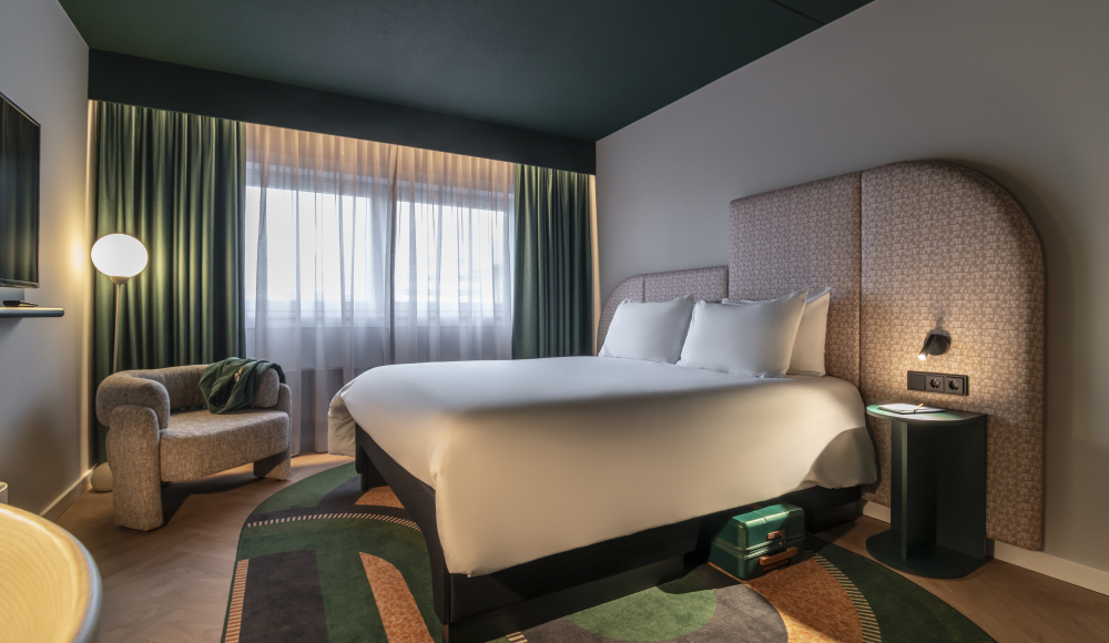 A stylish hotel room featuring a bed, an armchair, and windows with curtains.