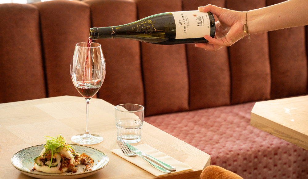 Eine Hand schenkt Rotwein in ein Glas neben einem Teller mit Essen.