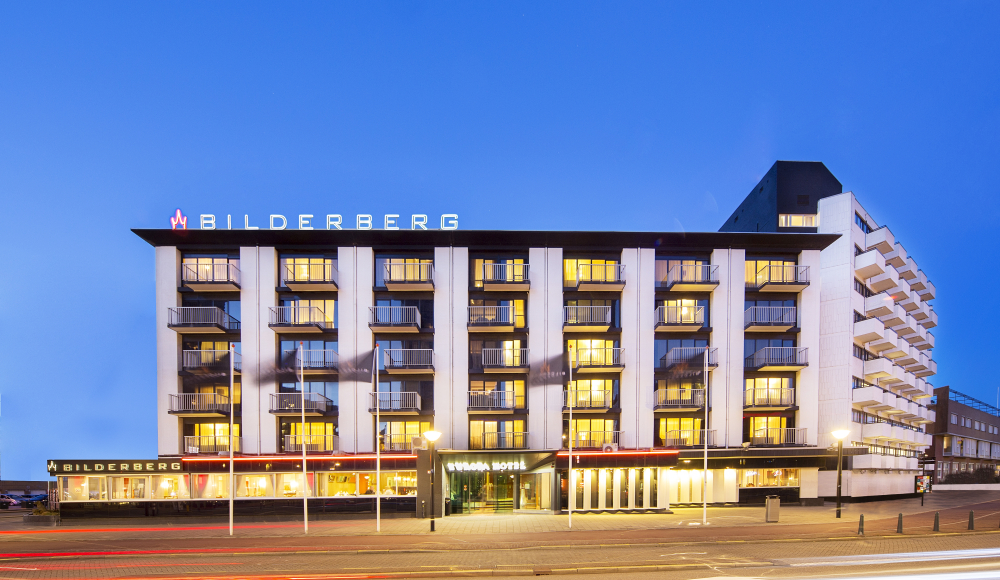 Foto van de voorkant van het hotel in de schemer met lichten aan. 