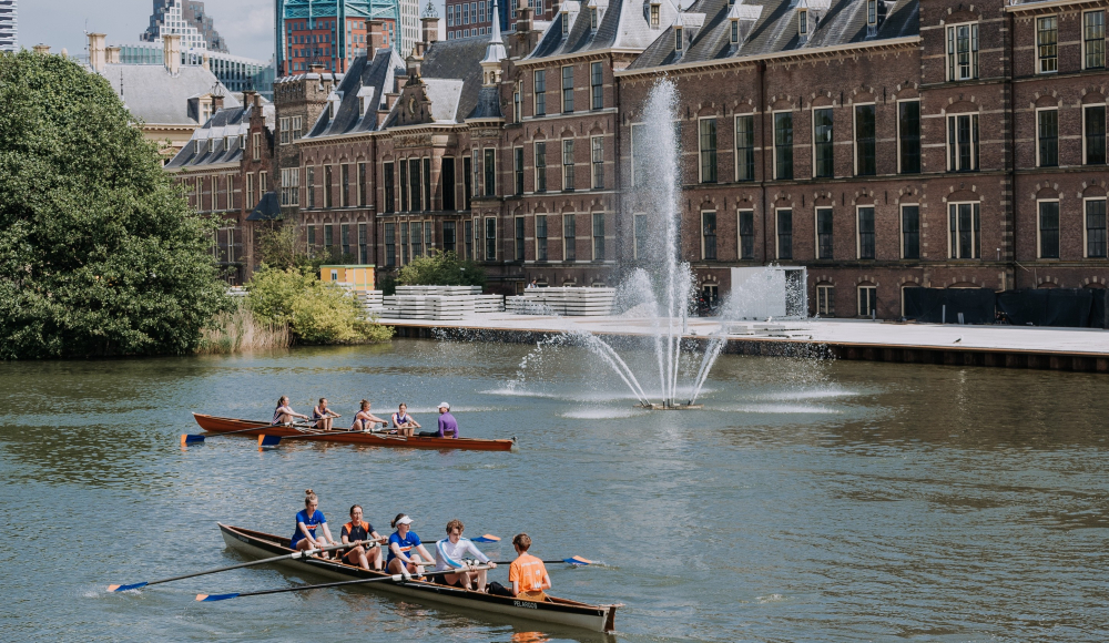 Two rowing boats with people on the Hofvijver, with a fountain and historic buildings in the background.
