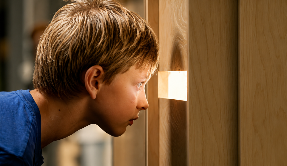 A boy curiously peering through a wooden slit.