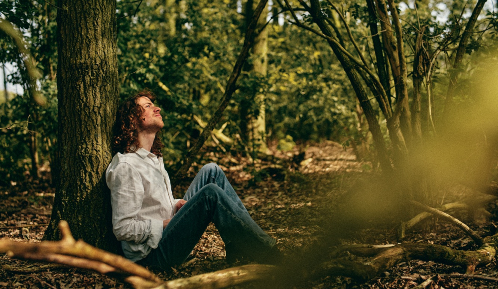 Ein junger Mann mit lockigem Haar lehnt an einem Baum im Wald.