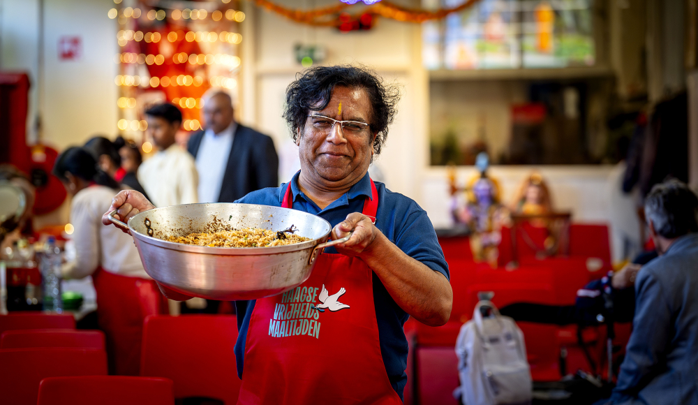 Een man in een rood schort houdt een pan met gekruid rijstgerecht vast in een sfeervolle, drukke ruimte.