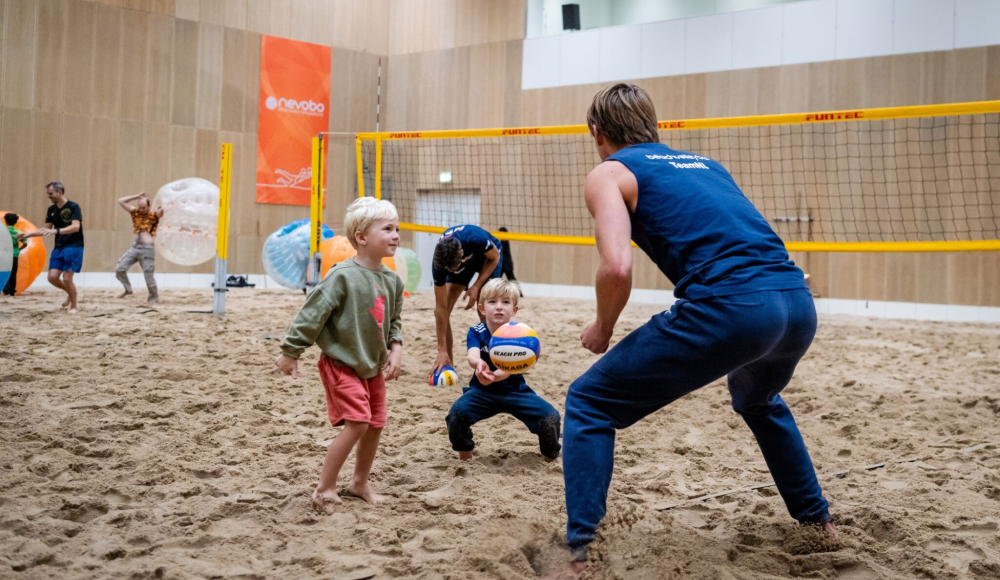 Twee kinderen spelen volleybal op een zandveld in een zaal.