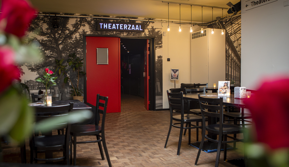 Vue de l'entrée d'une salle de théâtre avec une porte rouge et des tables.