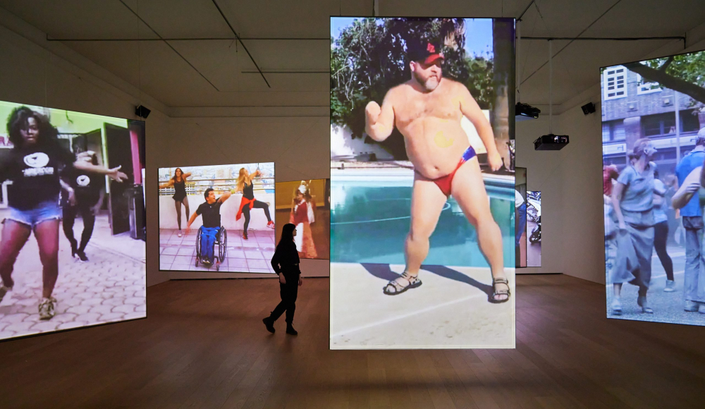Persoon in zwarte kleding loopt langs videoprojecties van dansende mensen.