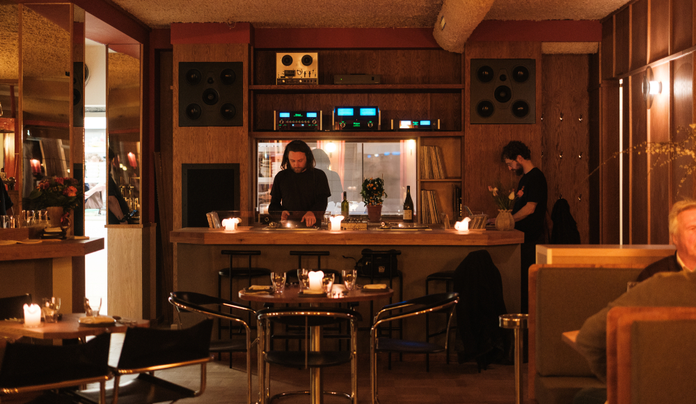 Twee mannen achter een houten bar met draaitafels en kaarslicht in een sfeervol retro interieur