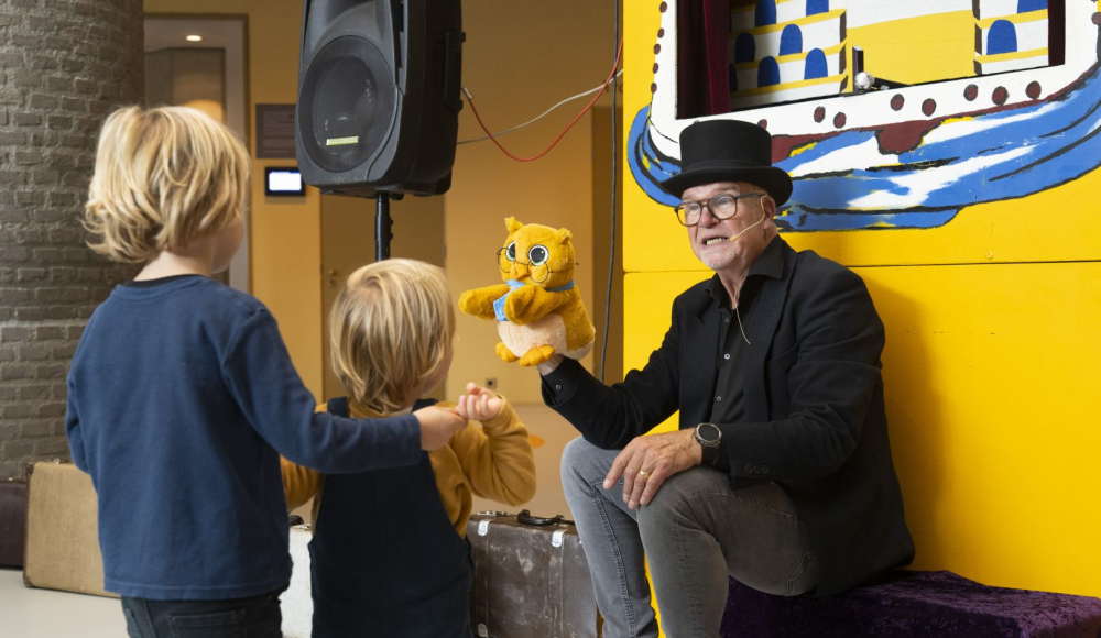 Een man met een hoed toont een handpop aan kinderen.