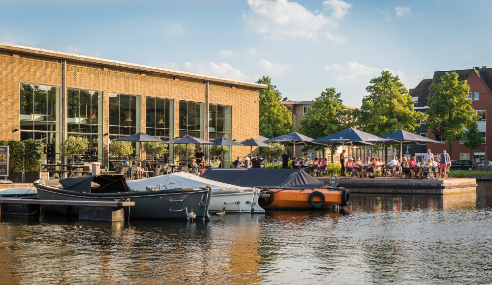 Terras vol met mensen aan de het water van de Fokkerkade met bootjes aan de kade.