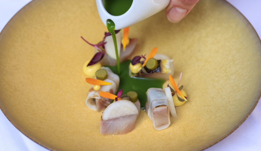 Une main verse une sauce verte sur un plat de poisson artistiquement présenté avec des garnitures.