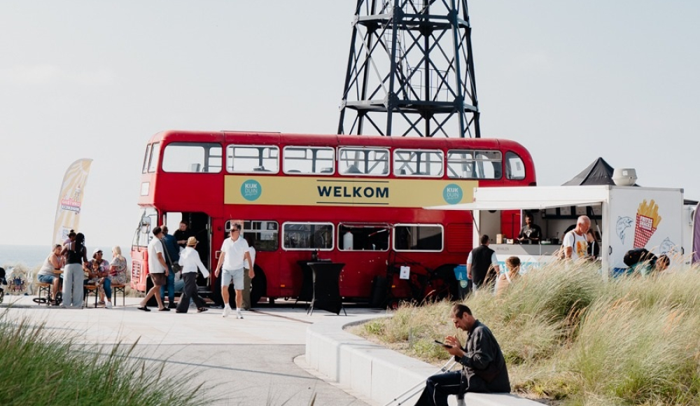 Een rode dubbeldeksbus met de tekst 'Welkom' naast een vuurtoren in Scheveningen.