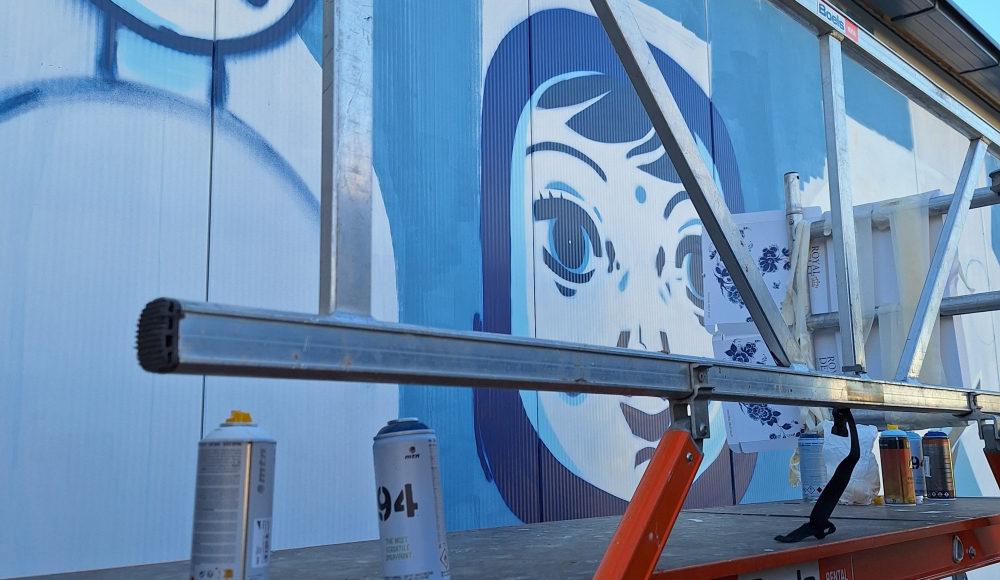 Een muurschildering van een gezicht in blauwe en witte kleuren, met verfspuitbussen op een ladder.