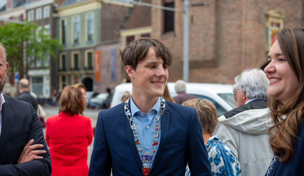 Een jonge man in een blauw jasje met een keten, lachend tussen een groep mensen. (c) Jelle Mes