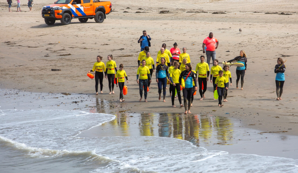 Safe@Sea. Een groep personen in surfkleding loopt langs het water.
