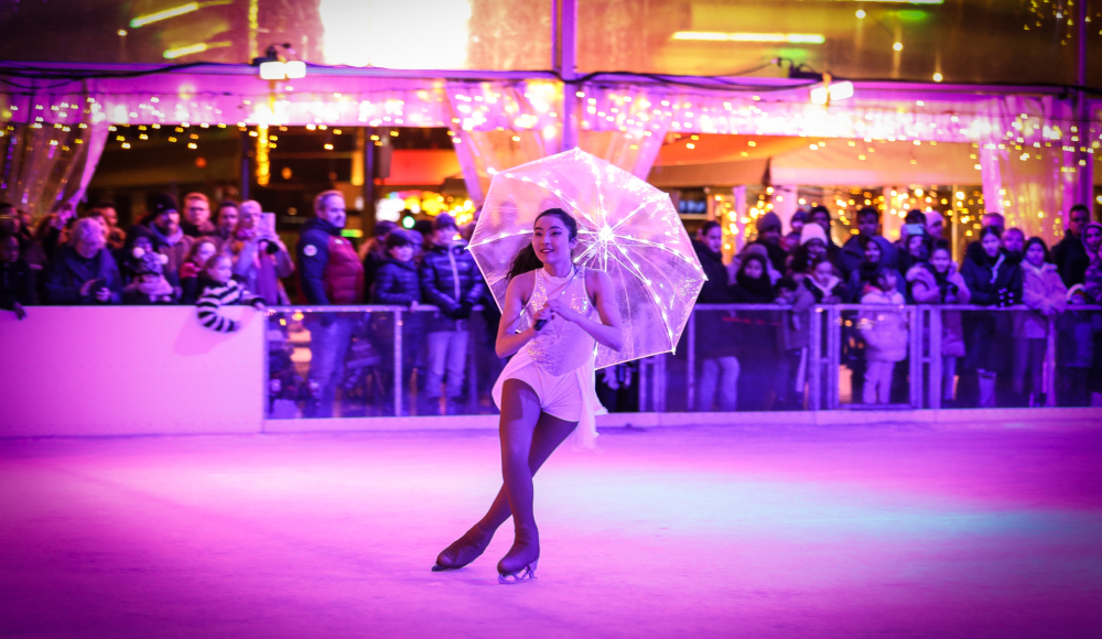 Eine Eiskunstläuferin in einem weißen Kostüm mit einem beleuchteten Regenschirm auf einer Eisbahn, umgeben von einer Menge.