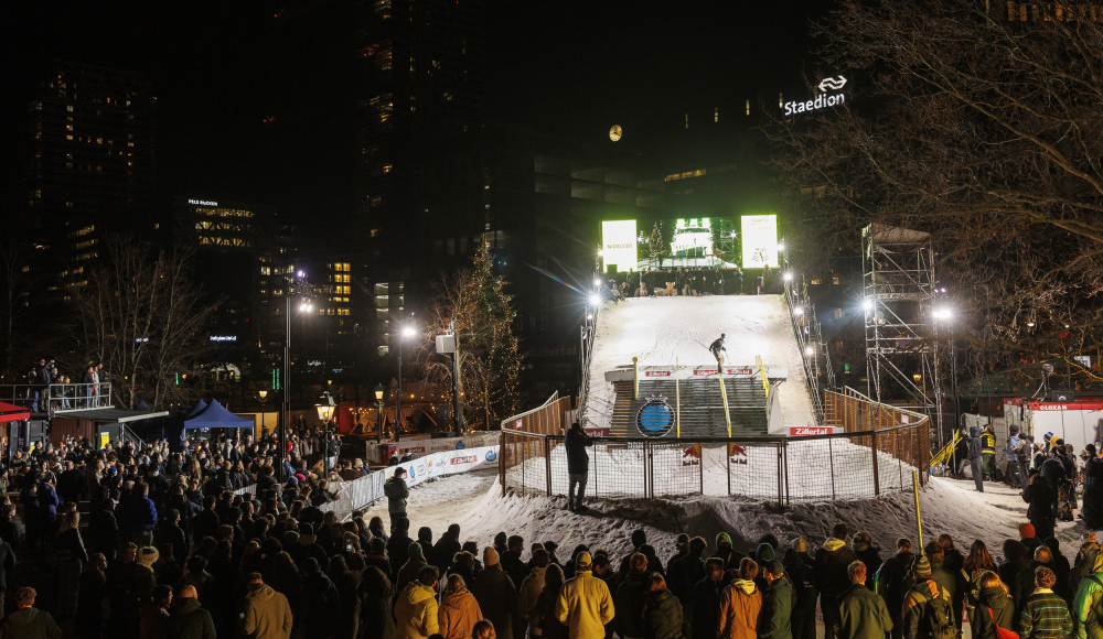 Événement de sports d'hiver en ville. Un snowboarder saute sur une pente, entouré de foules et de lumières de la ville.