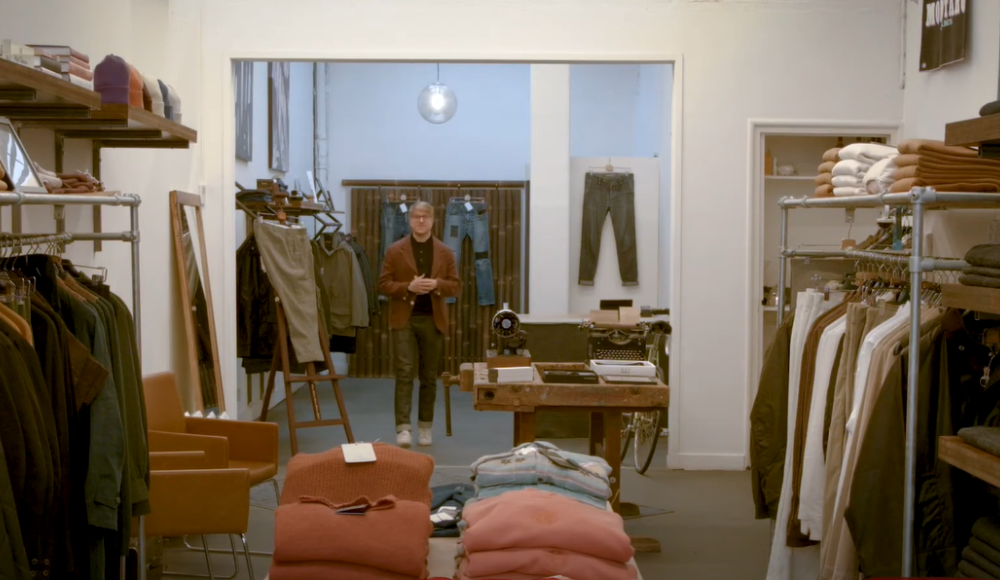 De eigenaar van kledingwinkel Store Du Nord poseert achterin zijn winkel. Aan weerszijden kledingrekken en in het midden tafels met truien.