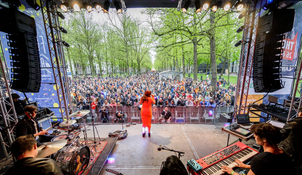 Zangeres op het podium op het Lange Voorhout tijdens The Life I Live. Ze is van achter gefotografeerd waardoor je het drukke publiek ziet.