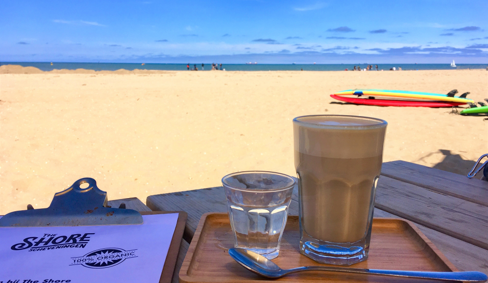 Groot glas koffie op een tafel met het strand en de zee op de achtergrond. 
