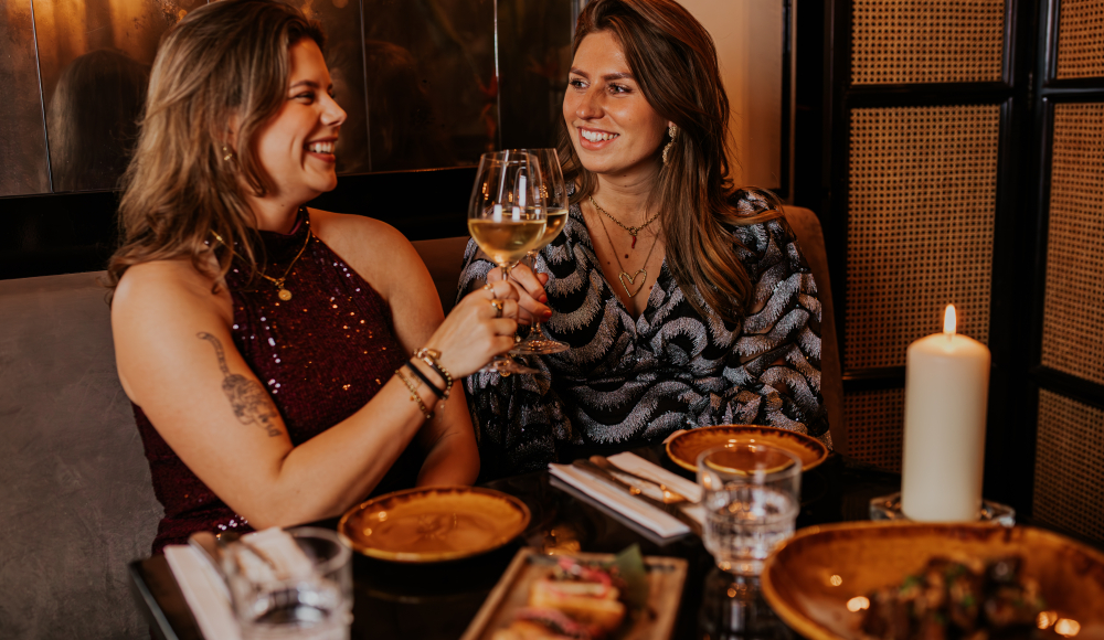 Two women toast with white wine at a table set with candlelight and plates of appetisers.