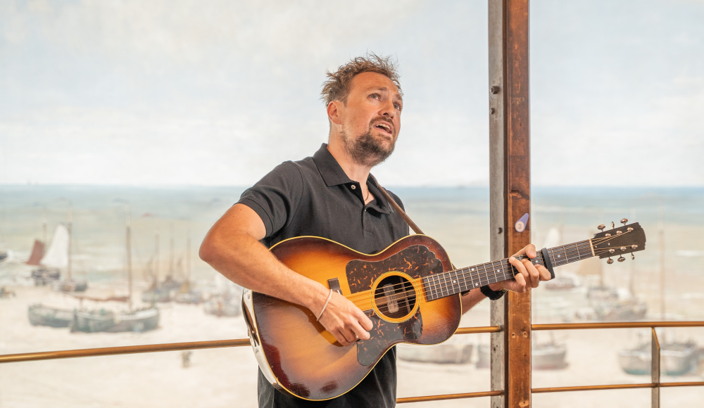 Tim Knol zingt en speelt gitaar met een zeezicht op de achtergrond.