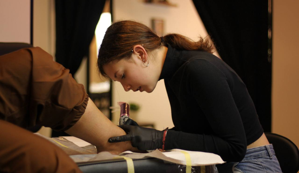 A young woman is tattooing a client's forearm.