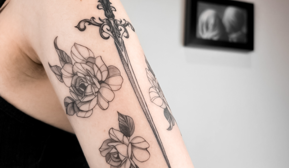 Black tattoo of flowers and a sword on an arm.