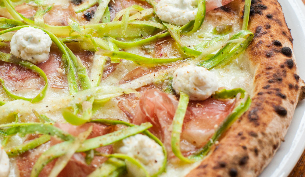 Close-up of a pizza topped with prosciutto, green vegetables, and cheese.