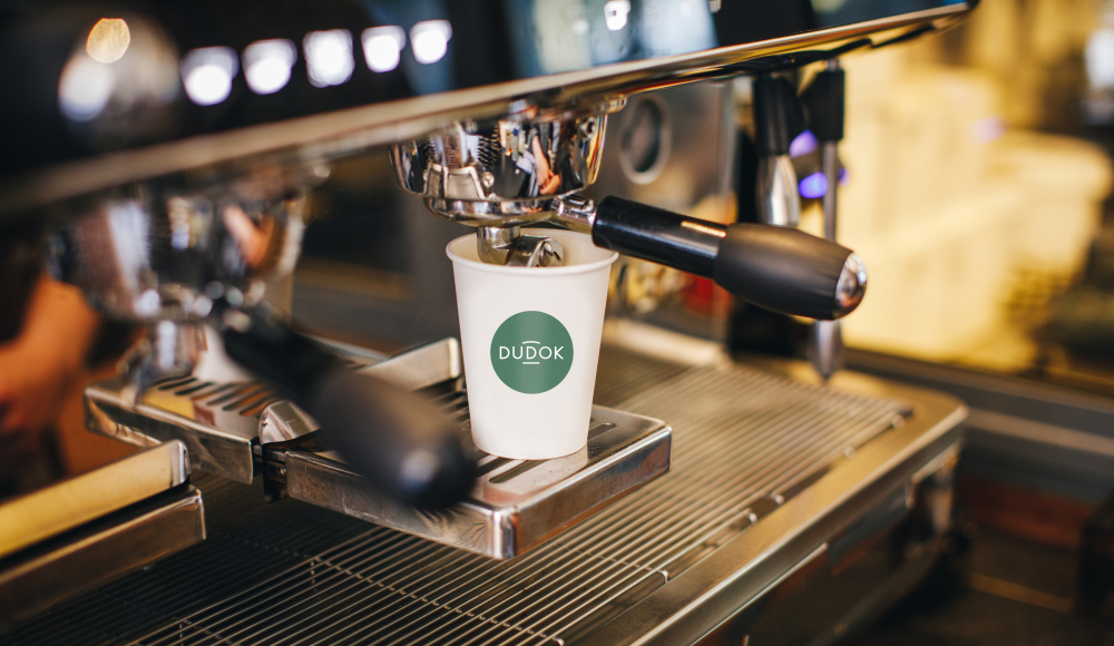 Kartonnen beker met logo van Dudok onder een professioneel koffieapparaat. 