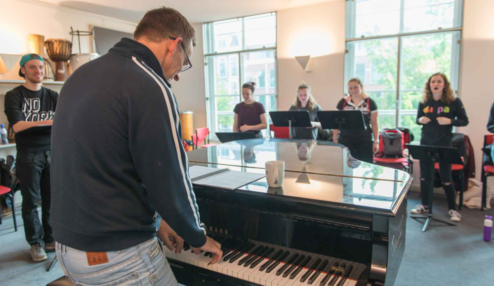 Man staand bij een piano die een koor begeleidt in het Koorenhuis 