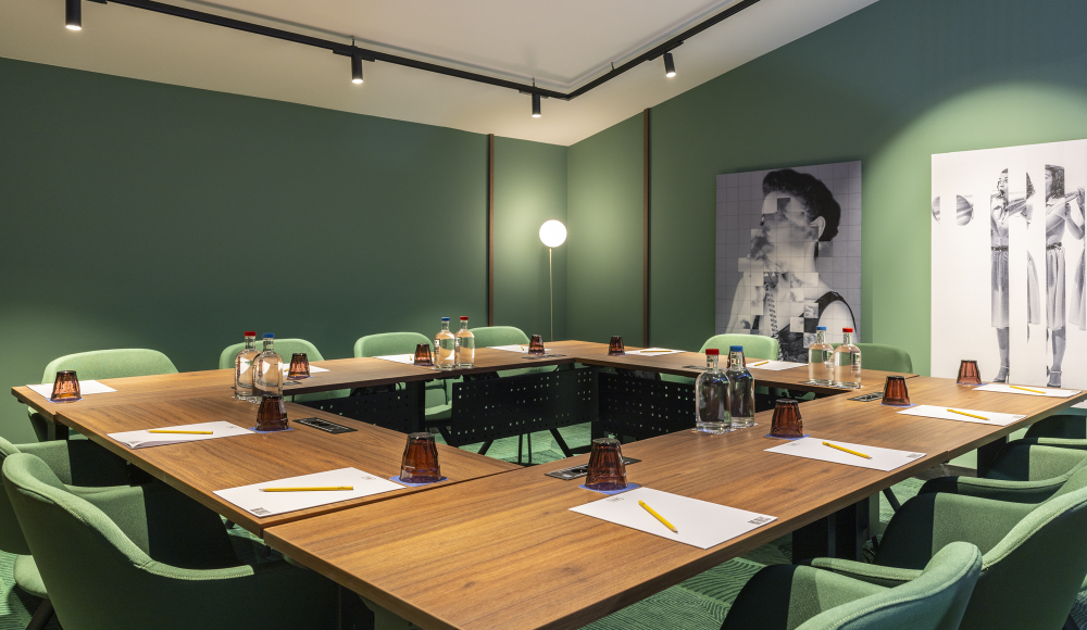 A meeting room with a wooden table, green chairs, water bottles, and notepads. Two artworks on the wall.