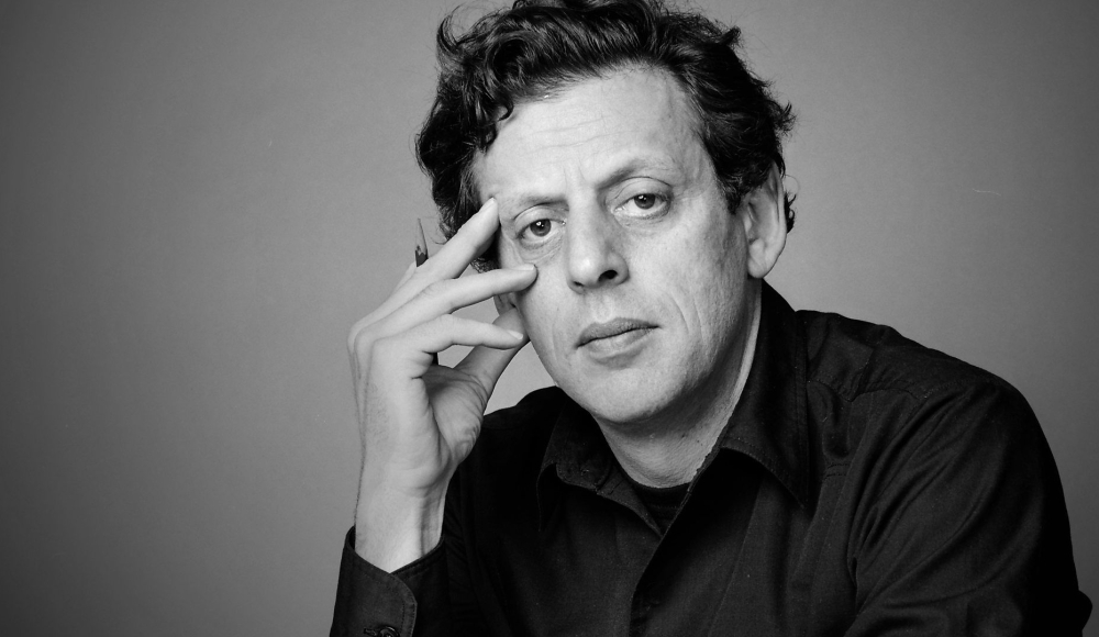 Zwart-witportret van een man met een intense blik, leunend op zijn hand, gekleed in een donker overhemd tegen een neutrale achtergrond.