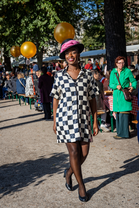 Een vrouw in een zwart-wit geruite jurk loopt met een glimlach op een evenement met gouden ballonnen op de achtergrond.