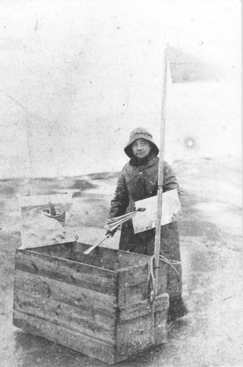 Een vrouw met een hoed bij een houten kist op het strand.
