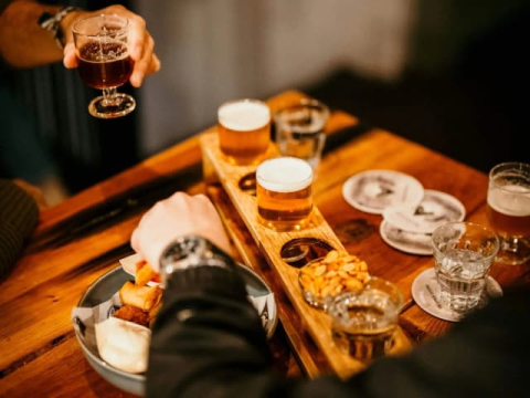Zwei Hände mit Getränken über einem Holztisch mit einer Bierprobe und einem Teller Snacks.