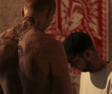 Tatoeages in een houten ruimte. Twee mannen met tatoeages, één vanuit de rug gezien.