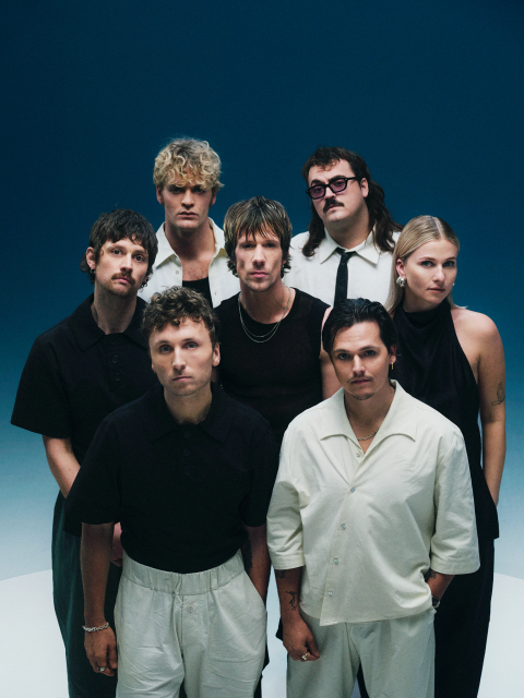 Zeven mensen staan in een groep voor een blauwe achtergrond.