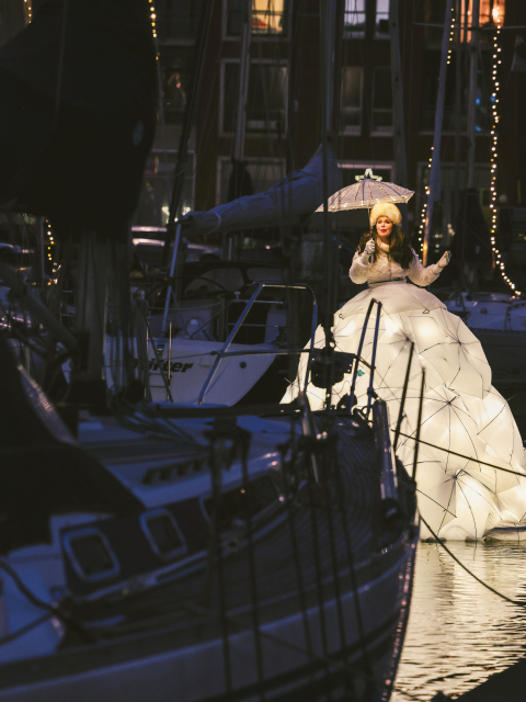 A singer in an extravagant winter costume with lights stands among the sailboats at Scheveningen.