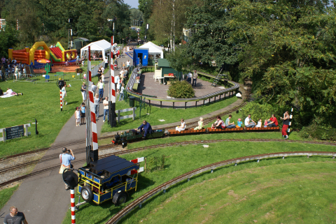 Modelstoomtreinenpark van bovenaf, midden in het groen van het Zuiderpark, met een rijdende trein vol kinderen en volwassenen, spoorovergangen en het station.